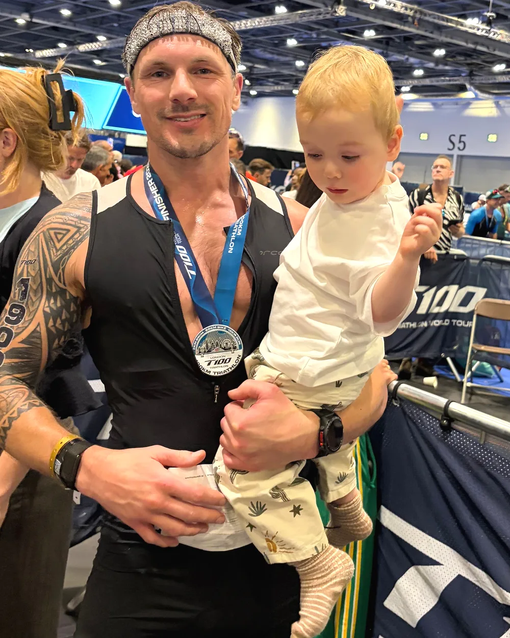 Jools Newsome holding his son at the finish of the T100 triathlon in London, wearing his finisher medal.