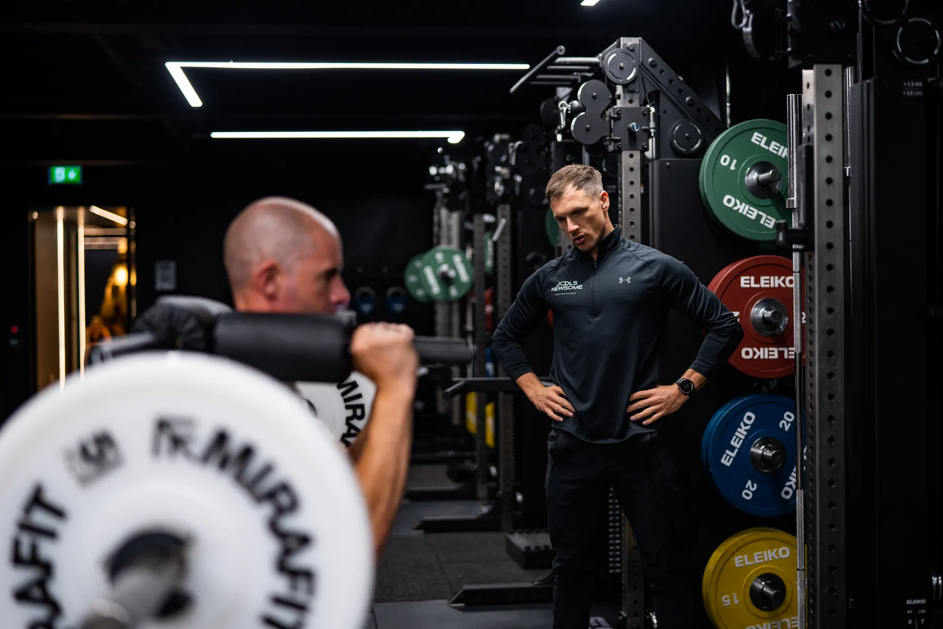 Jools Newsome on the training floor at UNTIL, hands on hips behind a loaded Eleiko barbell.